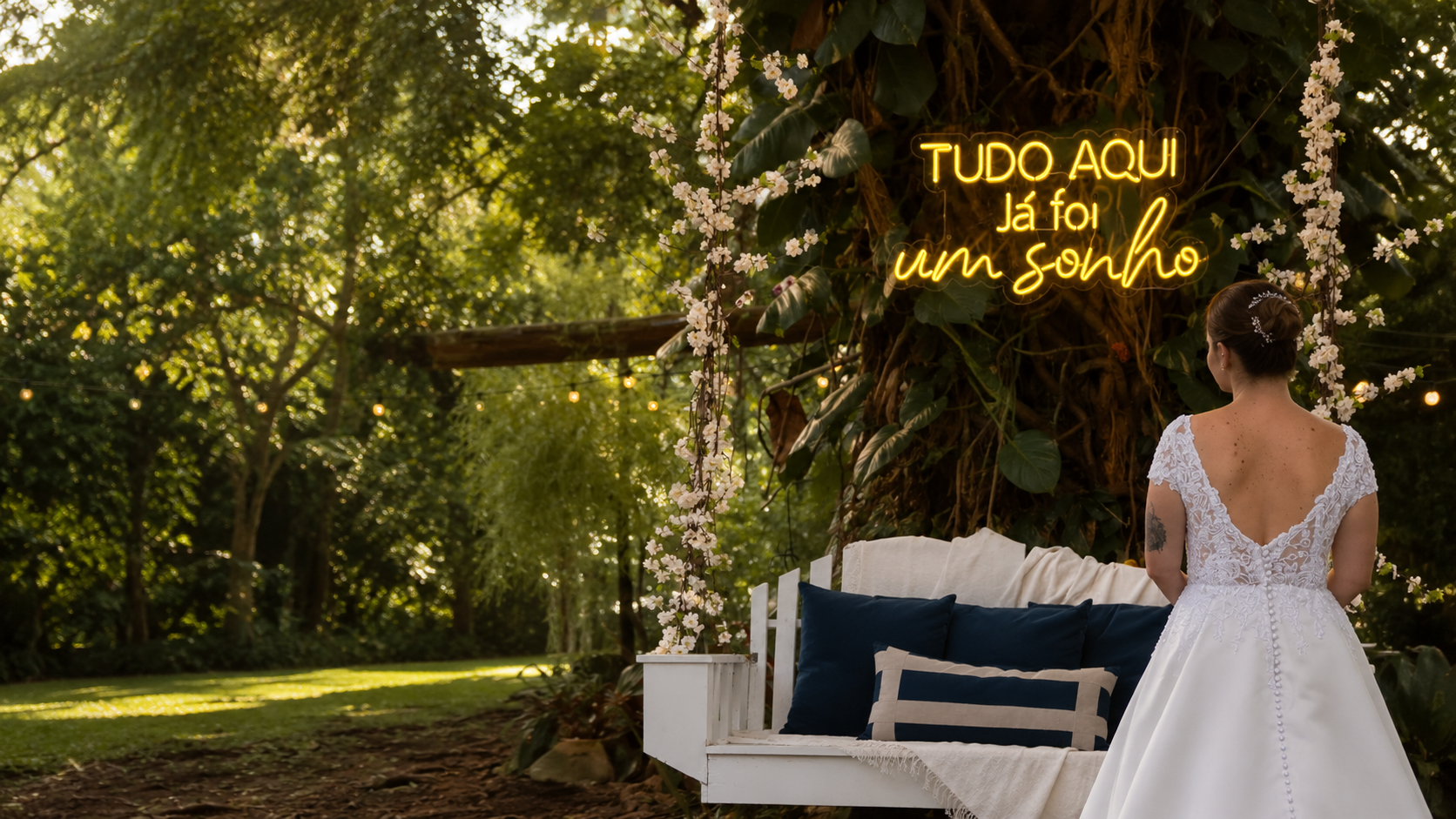 Noiva de costas no jardim olhando para o balanço com neon 'Tudo aqui já foi um sonho'